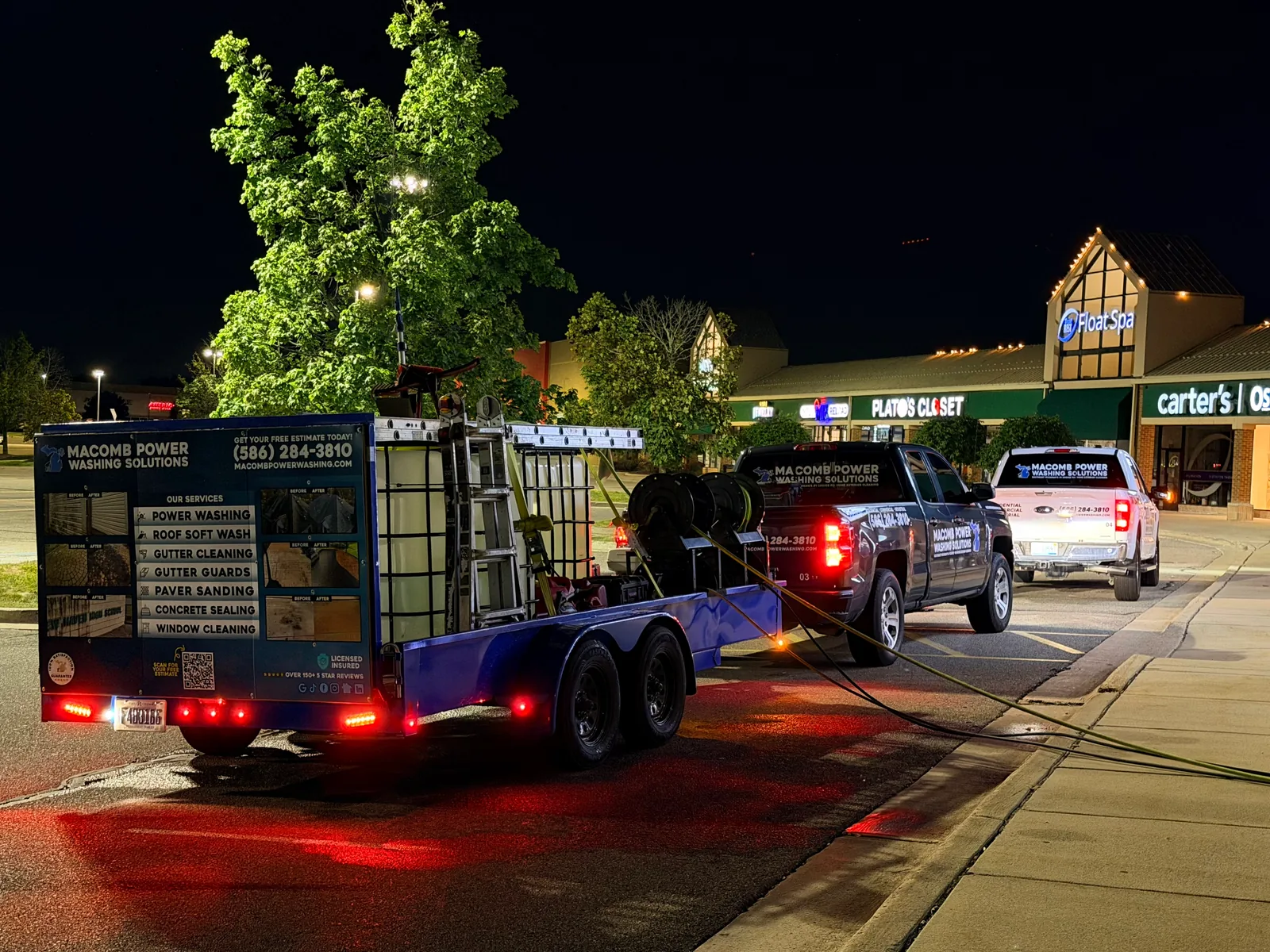 The Macomb Power Washing Solutions branded trailer and two service trucks staged at a shopping plaza at night, hoses run out across the wet lot