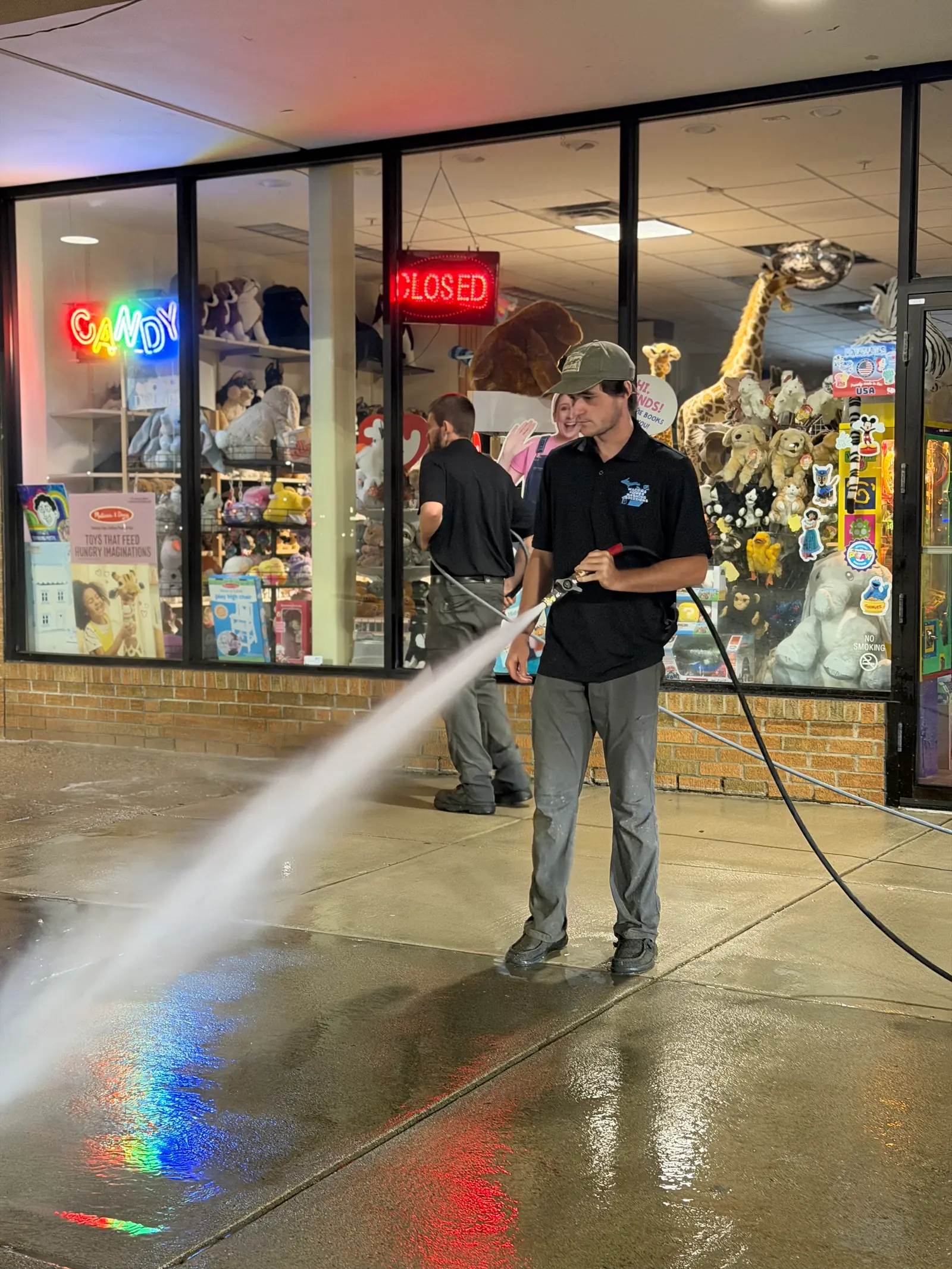 Two MPWS technicians washing a retail walkway after closing, storefront lights reflecting off the wet concrete